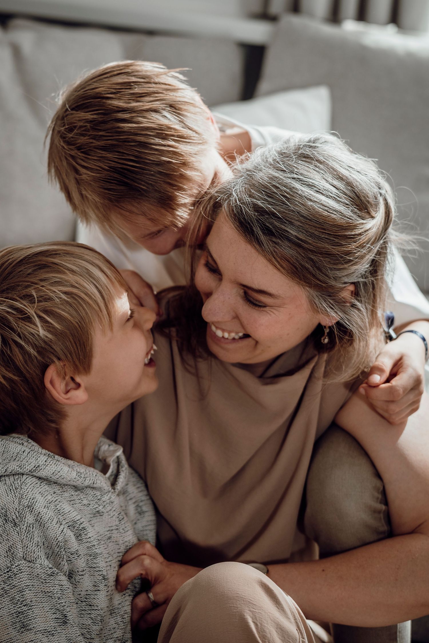 A smiling mother being embraced by two young boys on a cozy sofa at home.