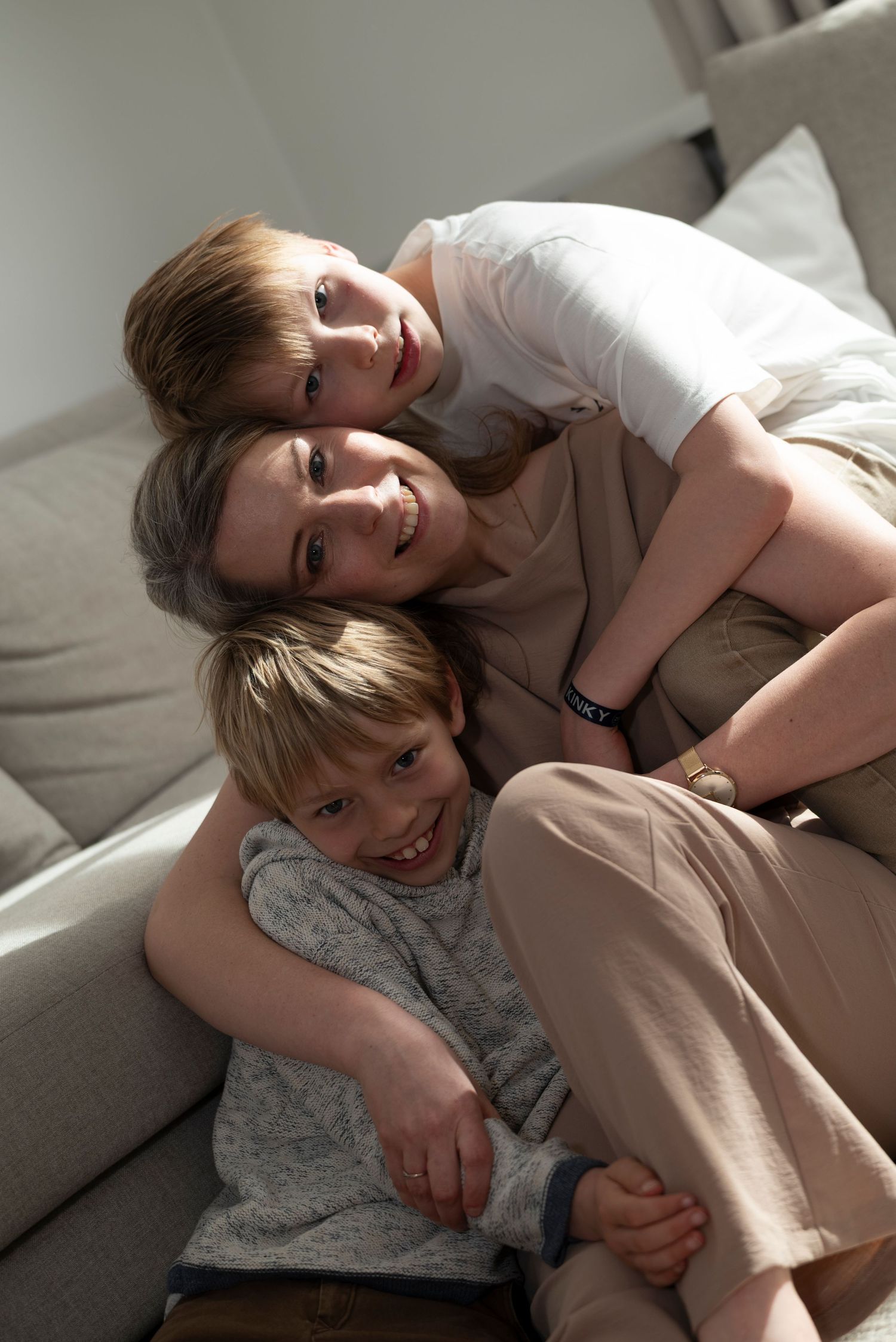 A mother and two young sons cuddling together on a grey sofa, smiling warmly.