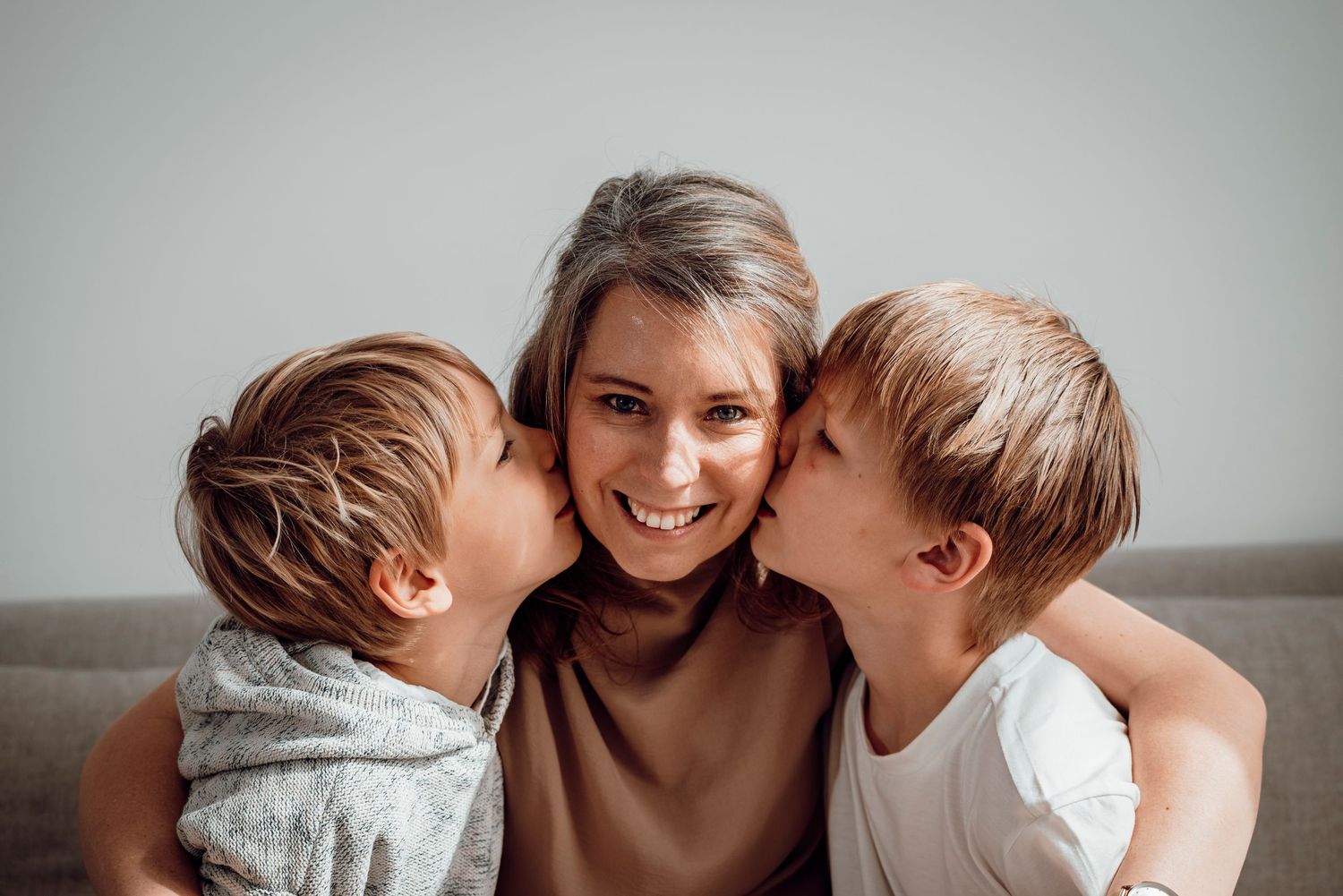 A happy mother receiving kisses on both cheeks from her two young sons on a sofa.