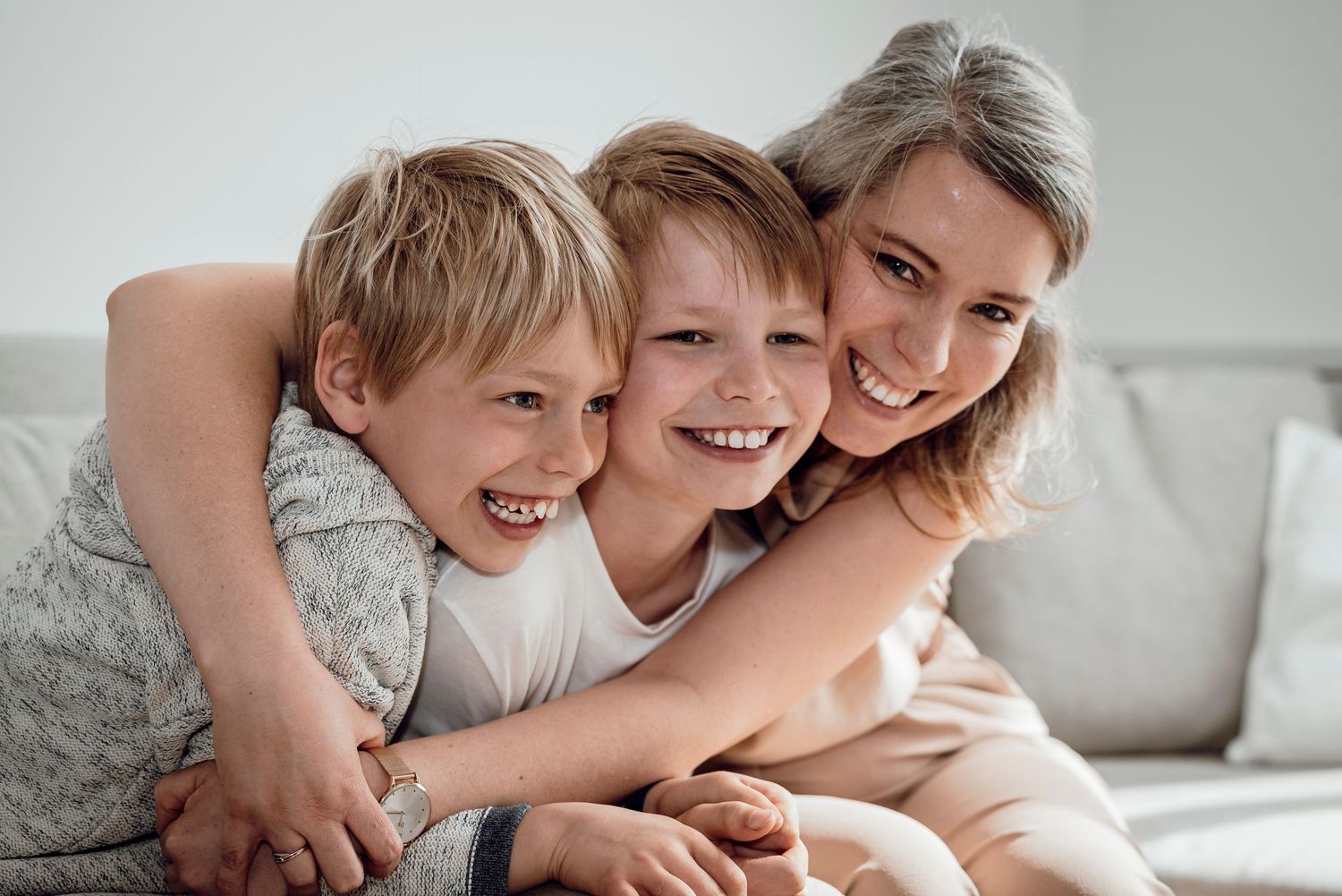 A joyful mother hugging her two laughing sons together on a light grey sofa.