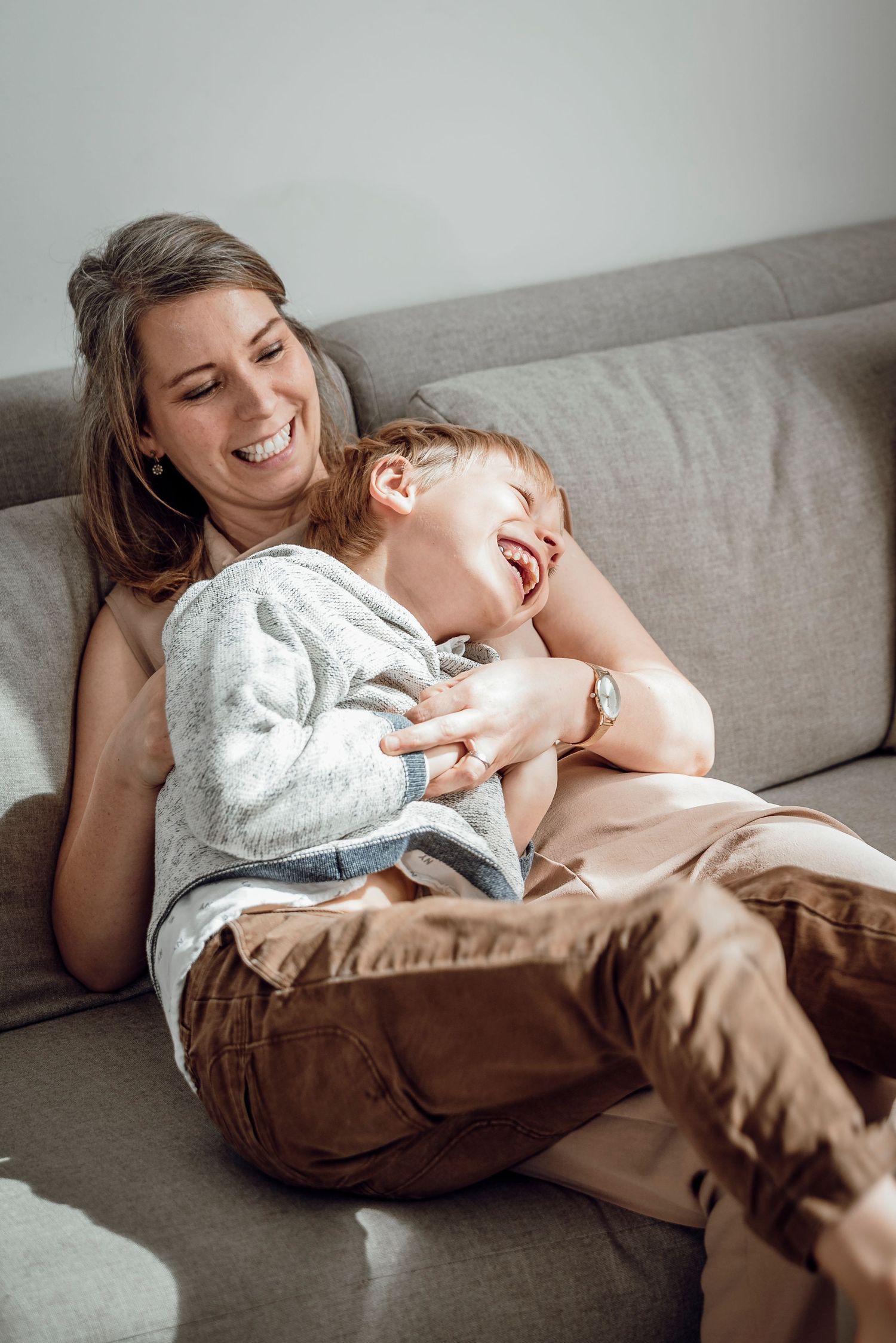 A laughing mother tickling her giggling young son on a grey couch at home.