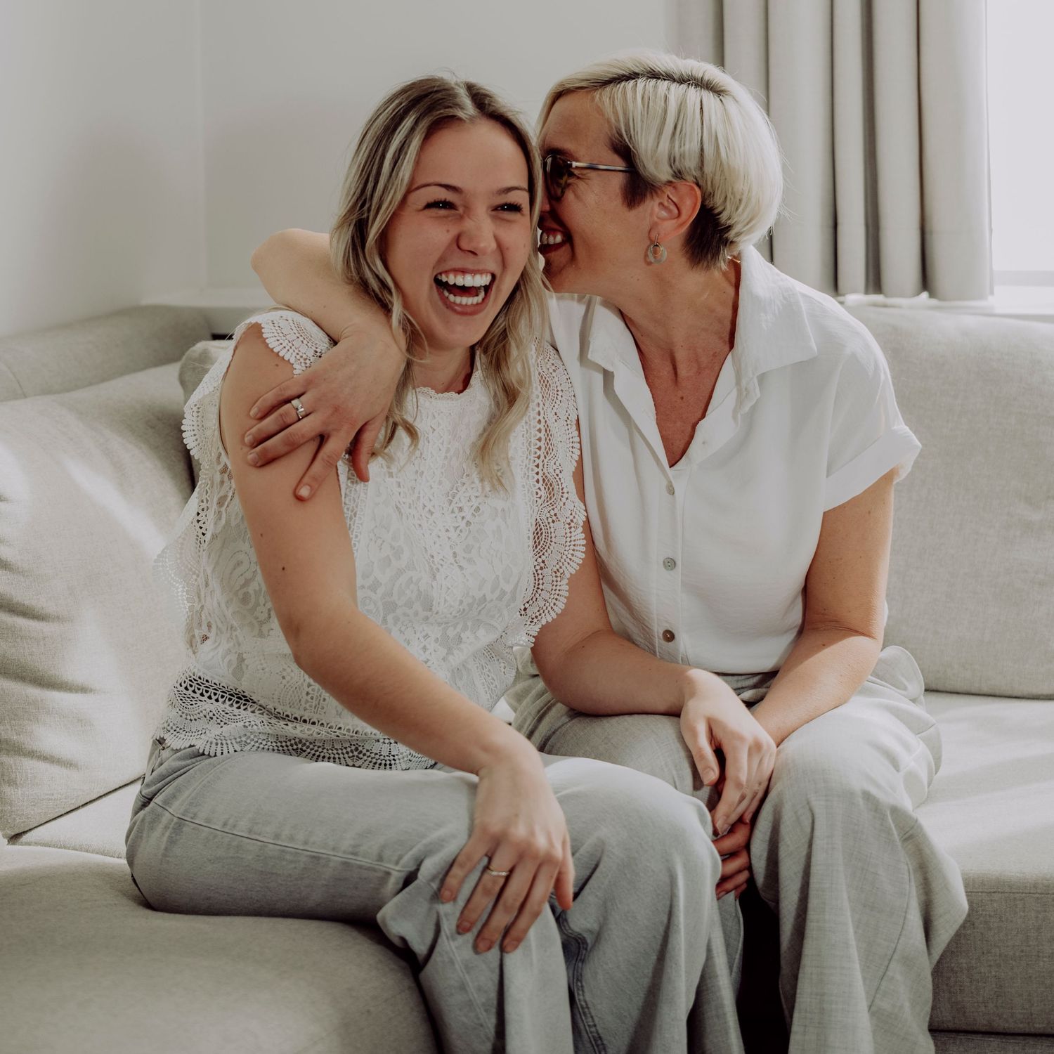 Two blonde women laugh joyfully together while sitting on a white couch in a bright, cozy living room.