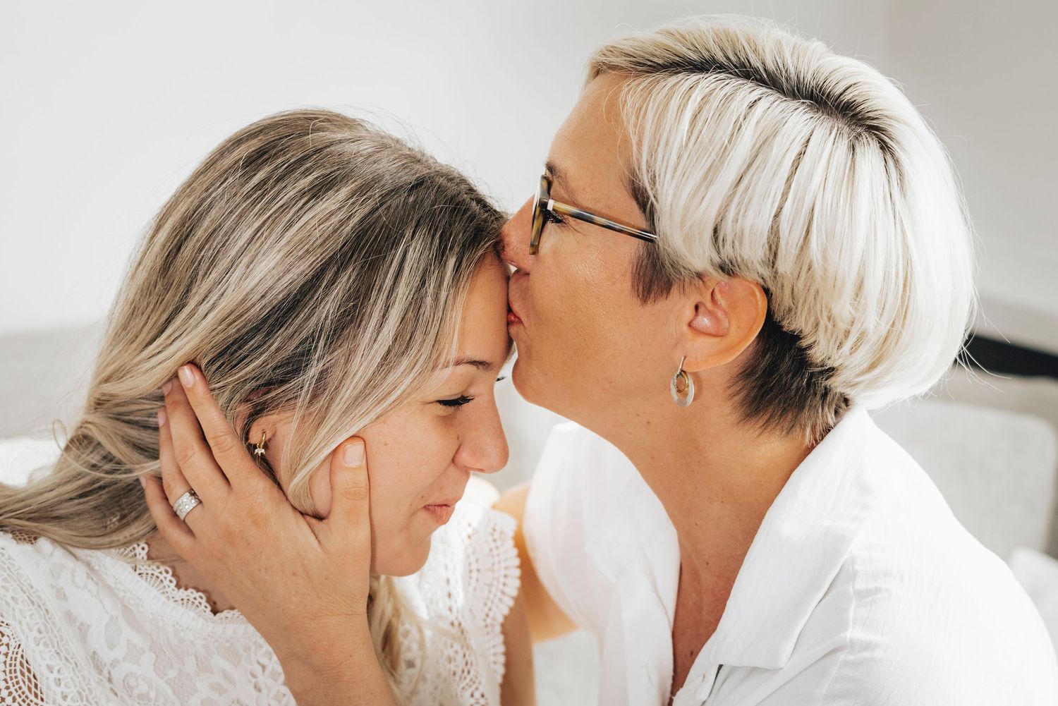 An older woman with short hair tenderly kisses the forehead of a younger blonde woman in a warm embrace.