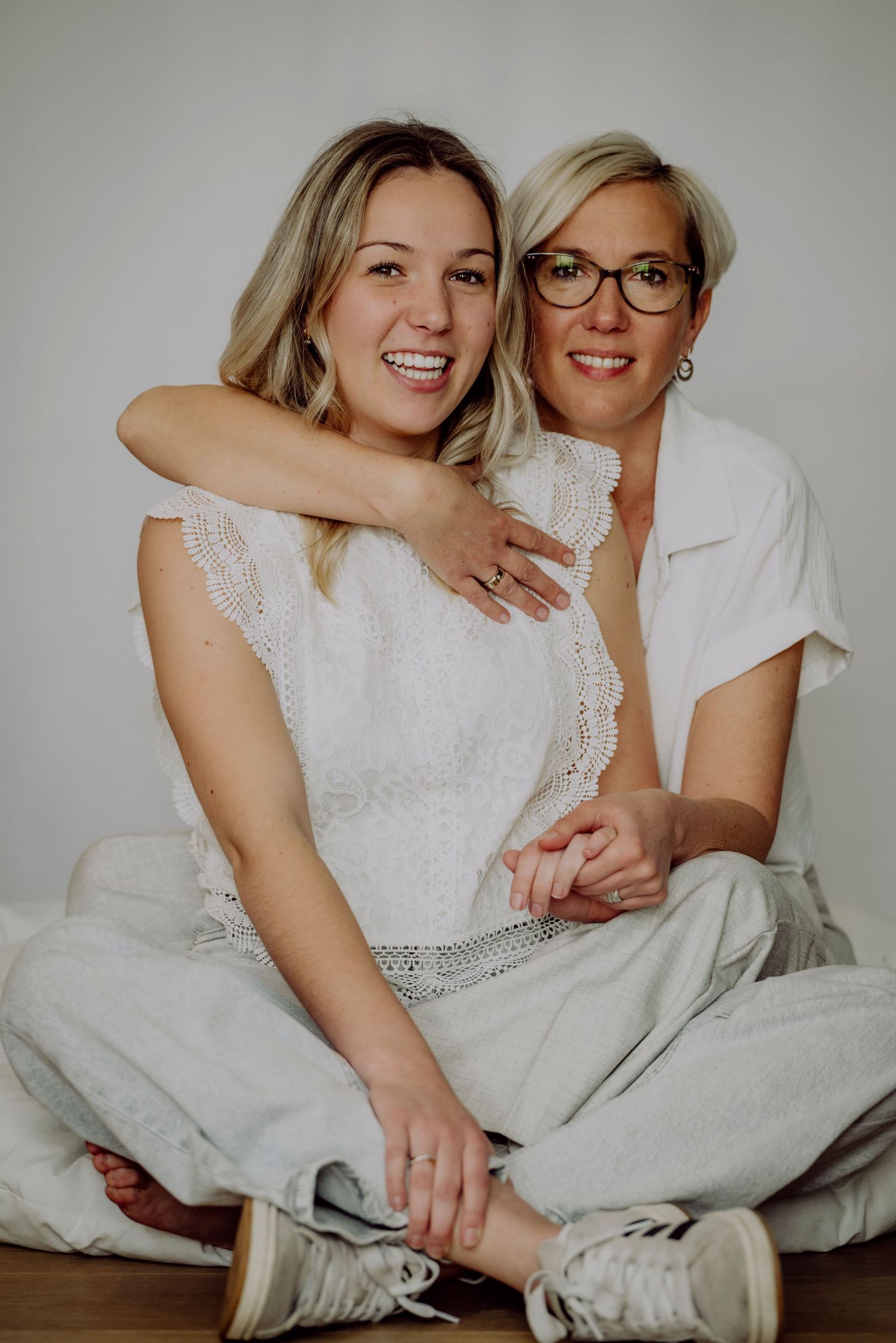 Two blonde women in white outfits sit closely together smiling warmly against a clean white background.