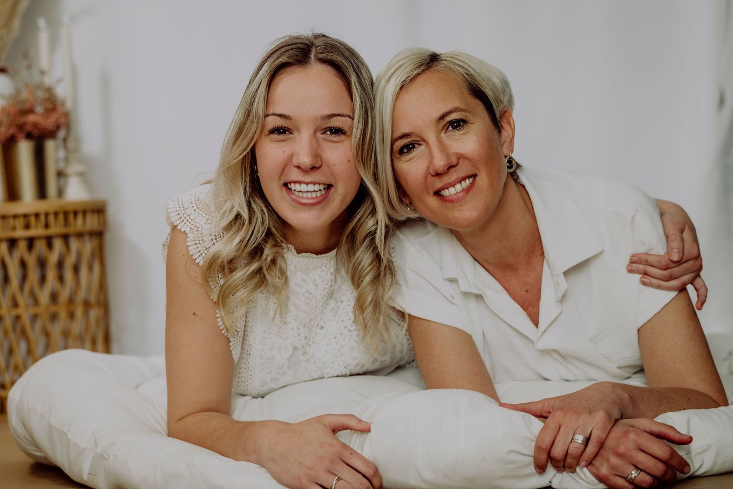 Two smiling blonde women in white outfits pose together lying down in a bright, cheerful indoor setting.