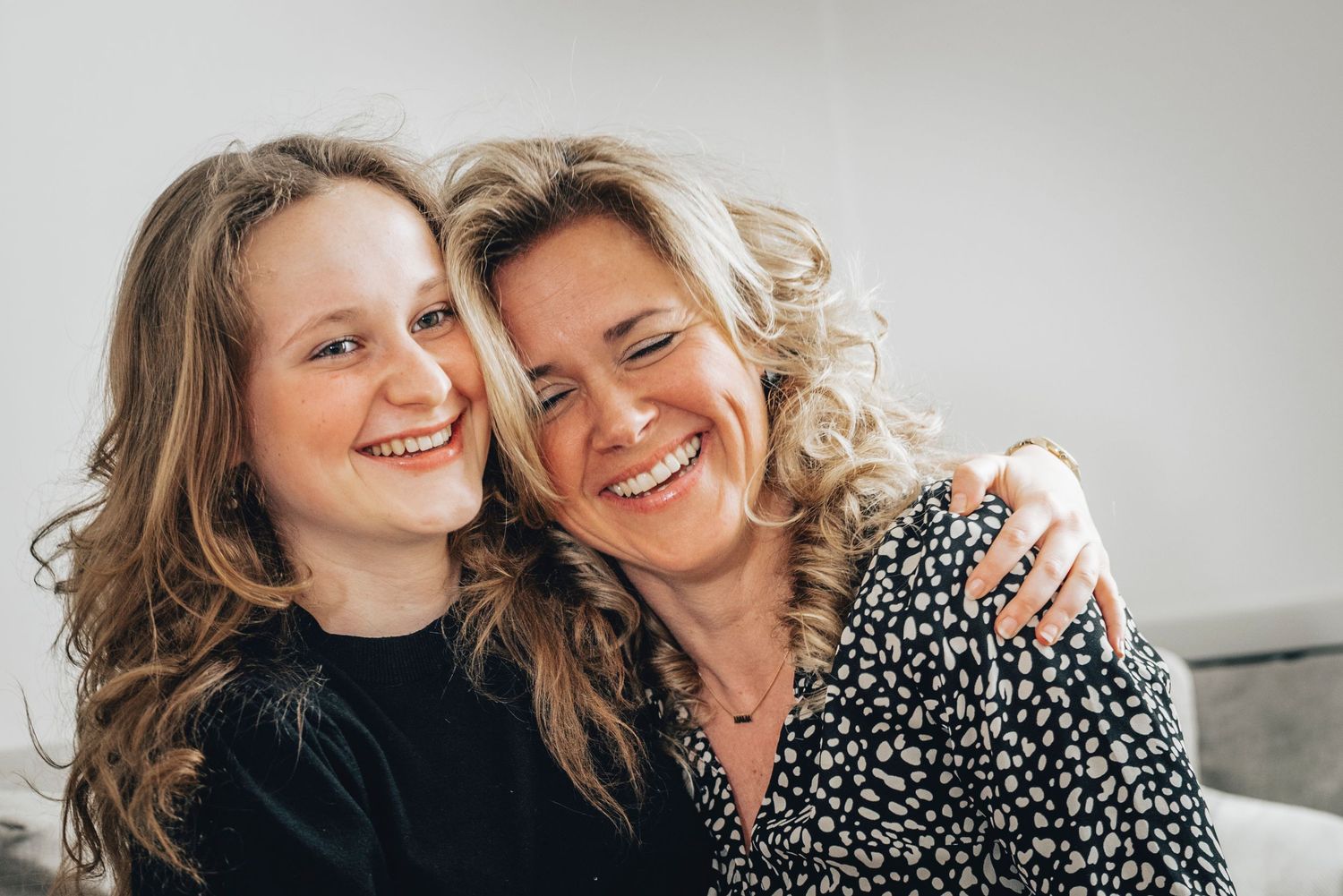 A young girl and a curly-haired blonde woman smile and embrace warmly while sitting together indoors.