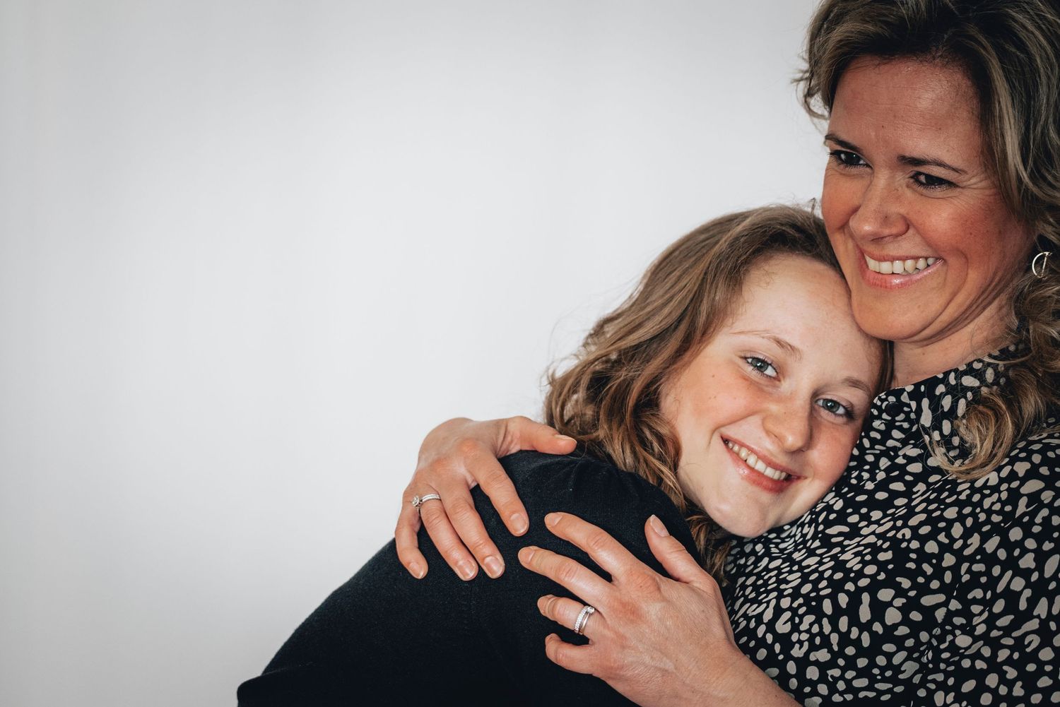 A woman hugging a smiling young girl from behind in a warm portrait photo.