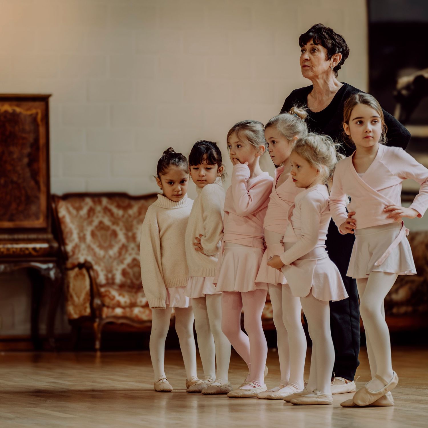 Ballet teacher guiding a group of young girls in pink outfits at barre.