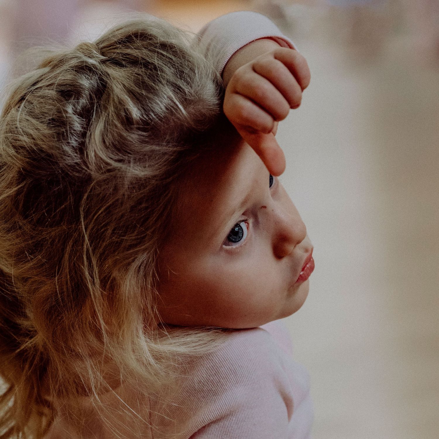 Curious toddler with curly hair resting hand on forehead, looking sideways with blue eyes.