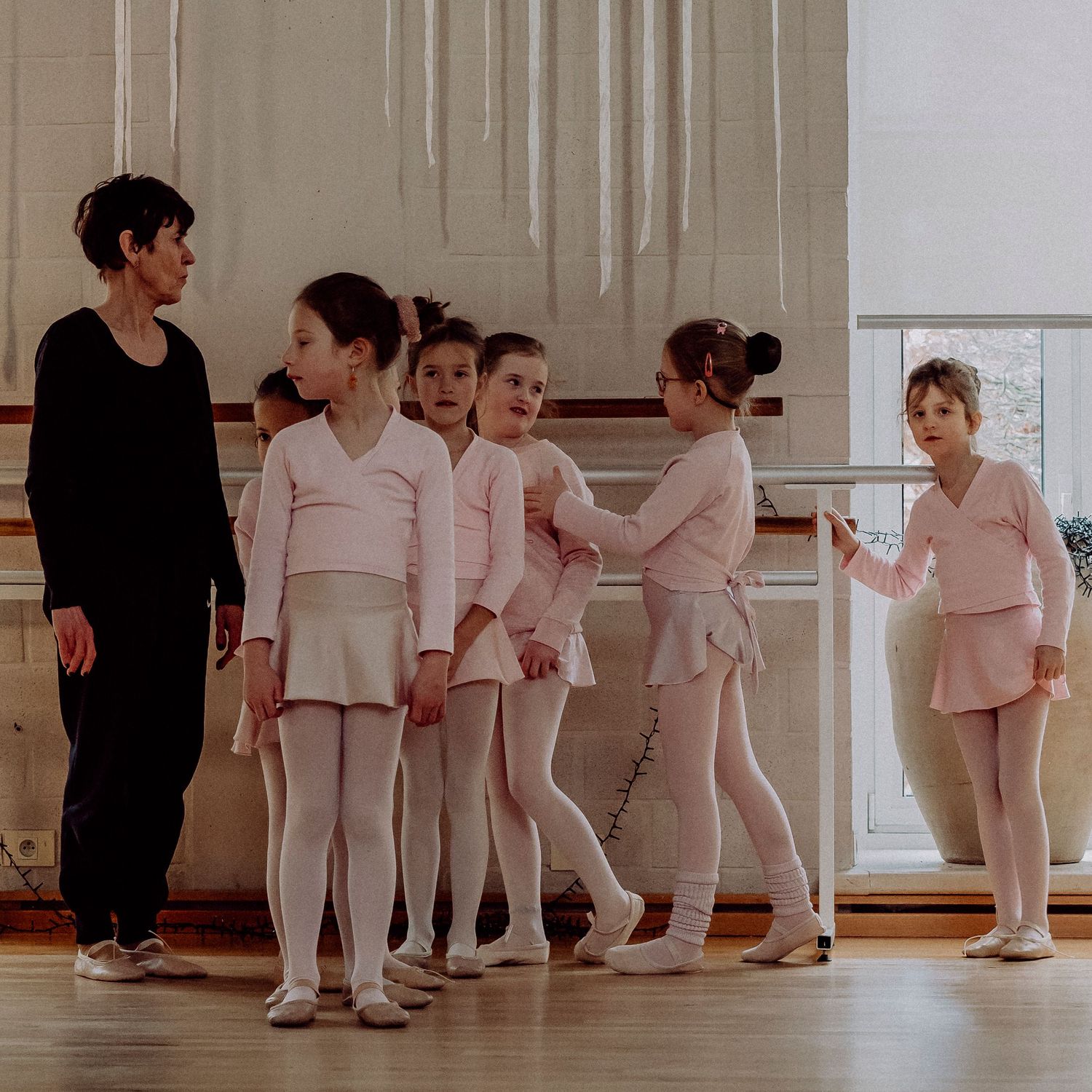 Ballet teacher instructing young girls in pink leotards at studio barre.