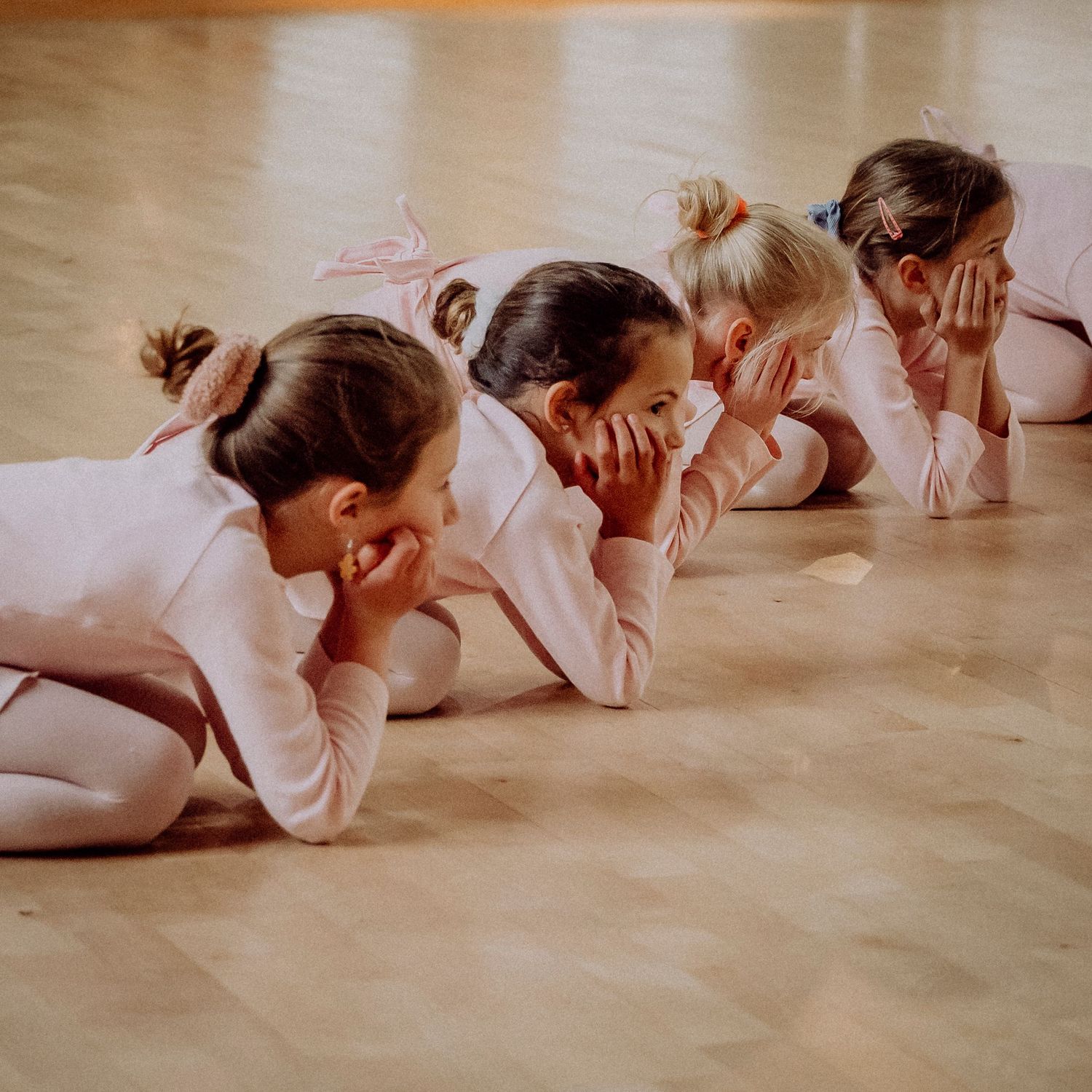 Four young ballet students in pink leotards lying on studio floor, smiling and giggling together.