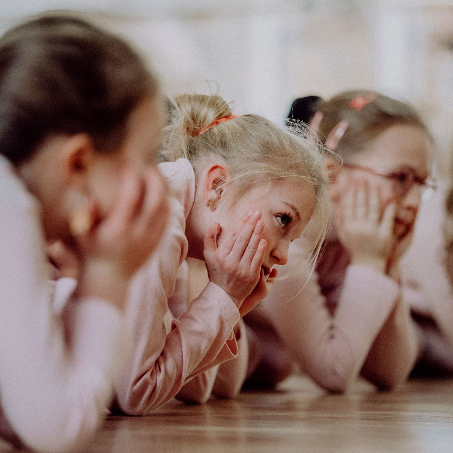 Young ballet girls in pink leotards lying on wooden dance studio floor, laughing and covering faces.