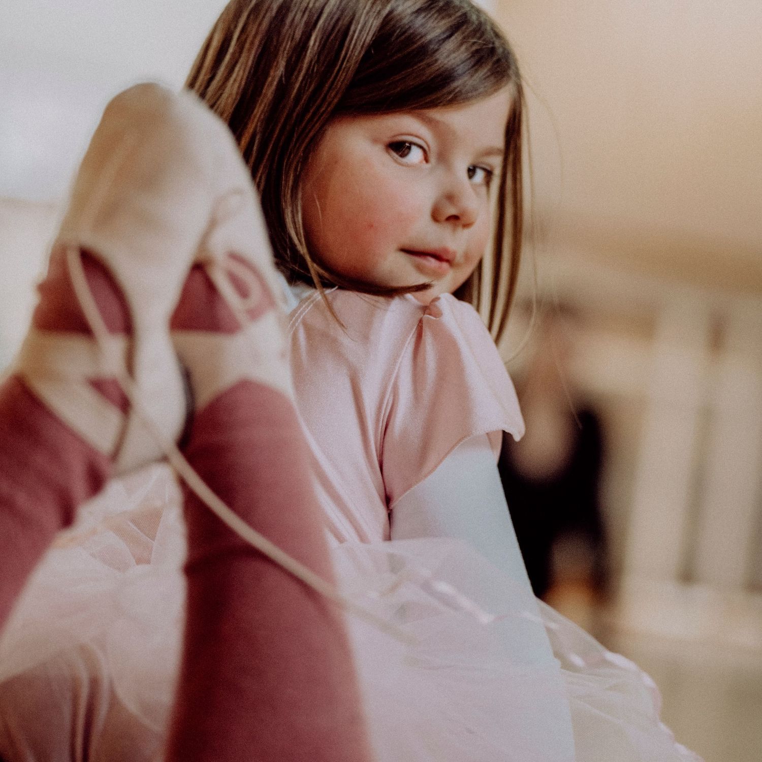 Young brunette ballet student in pink leotard holding pointe shoes, looking thoughtfully at camera.