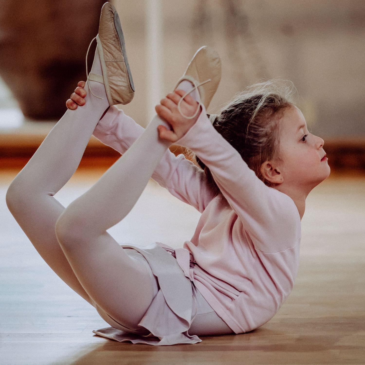 Young ballet student in pink leotard performing a bow pose stretch on wooden studio floor.