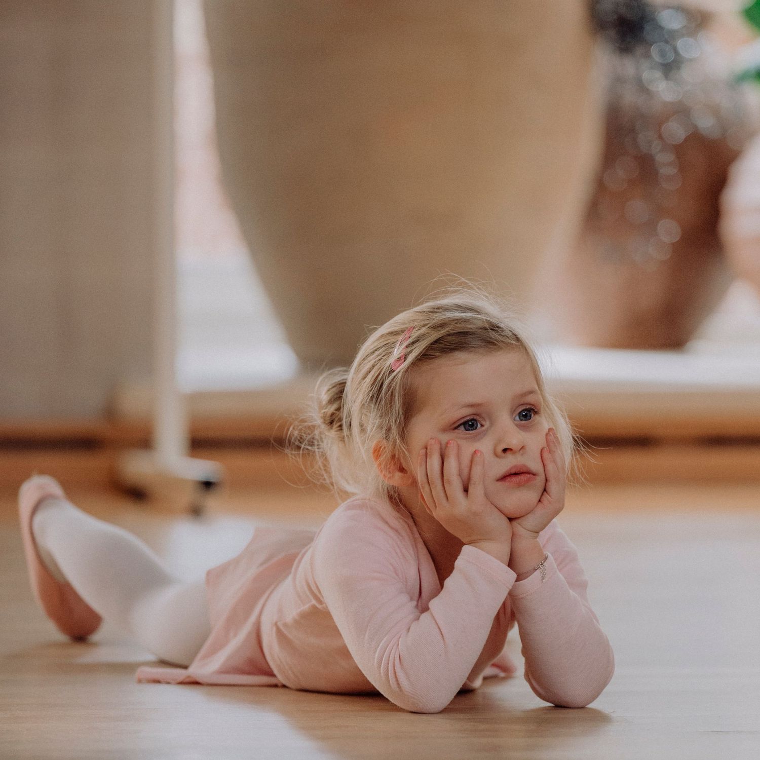 Little blonde ballerina in pink leotard lying on wooden floor, resting chin on hands with dreamy expression.