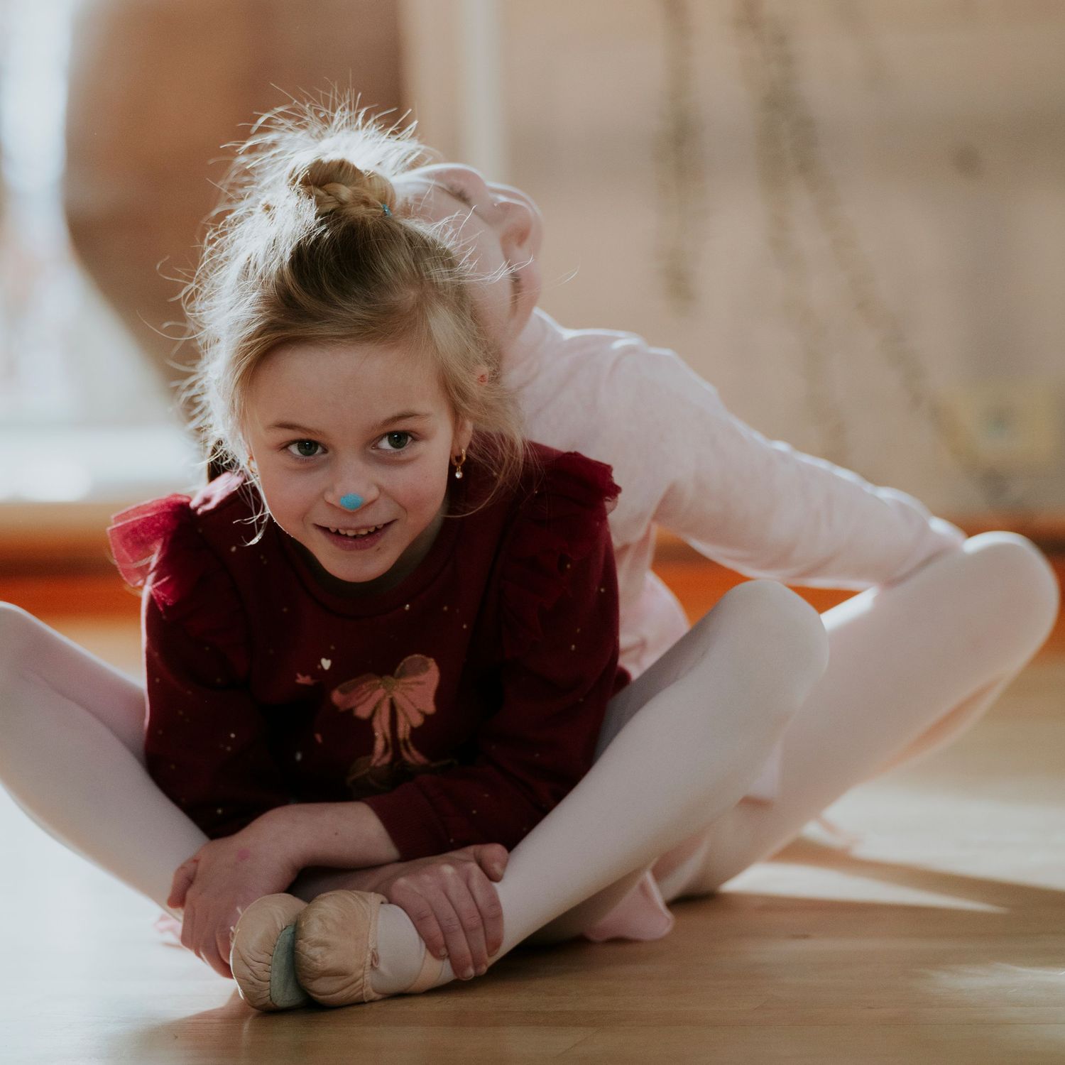 Smiling young girl in red velvet top and white tights stretching on ballet studio floor.