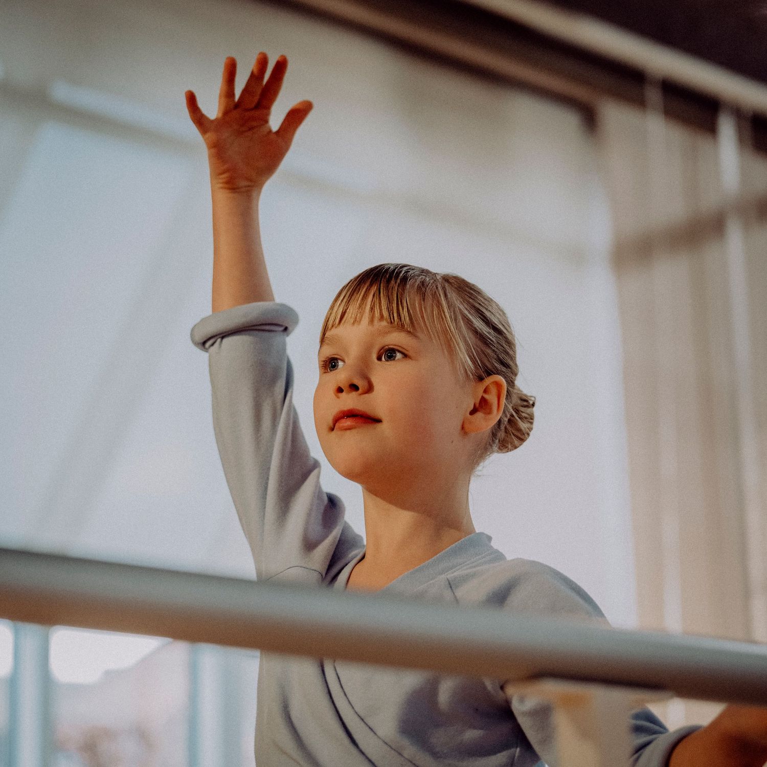 Young ballet student in blue leotard gracefully raising arm at barre in bright dance studio.