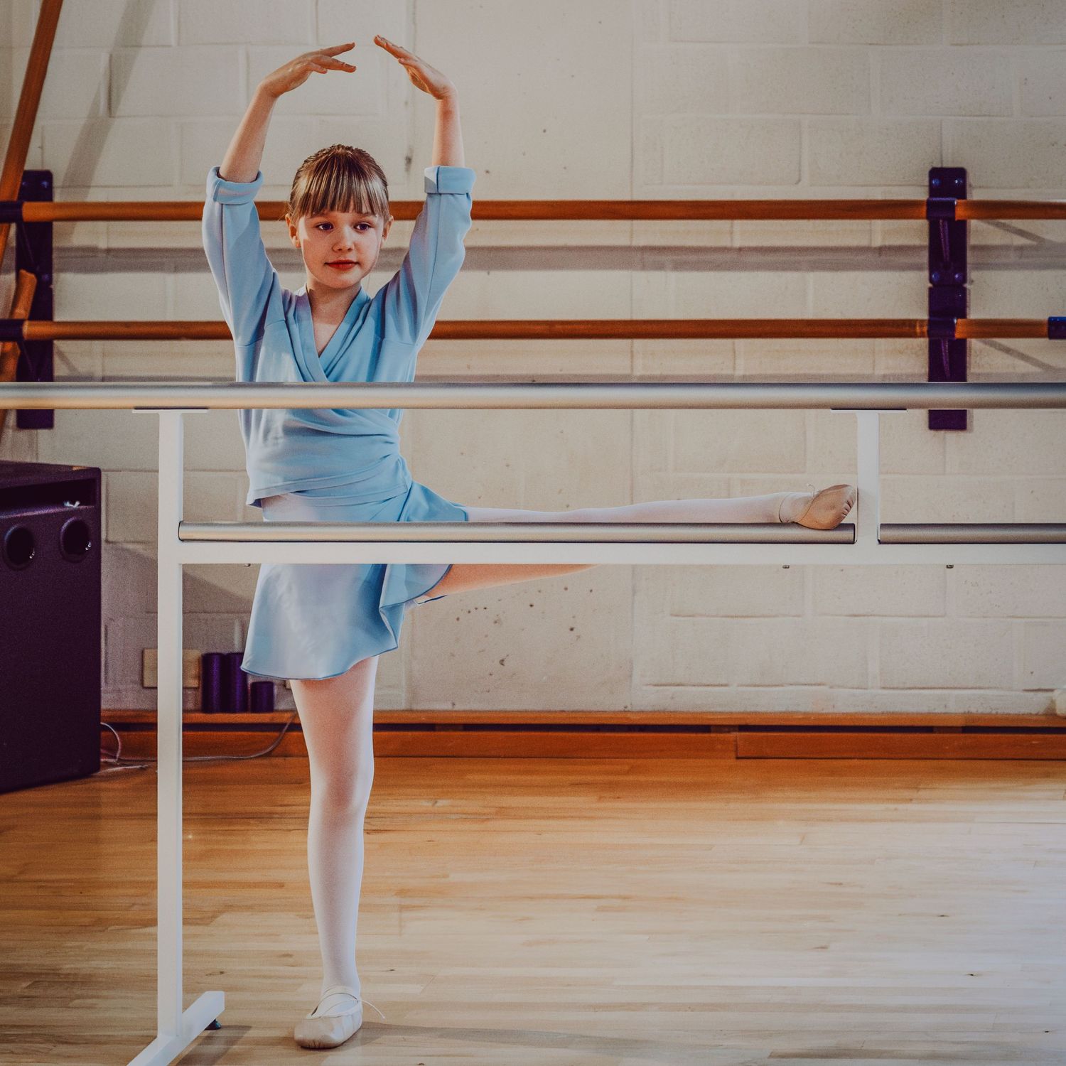 Young ballerina in blue leotard practicing leg extension at ballet barre in dance studio.