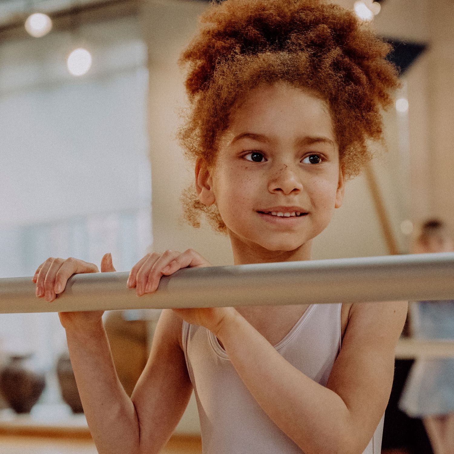Smiling young girl with curly hair holding ballet barre in white leotard during dance class.