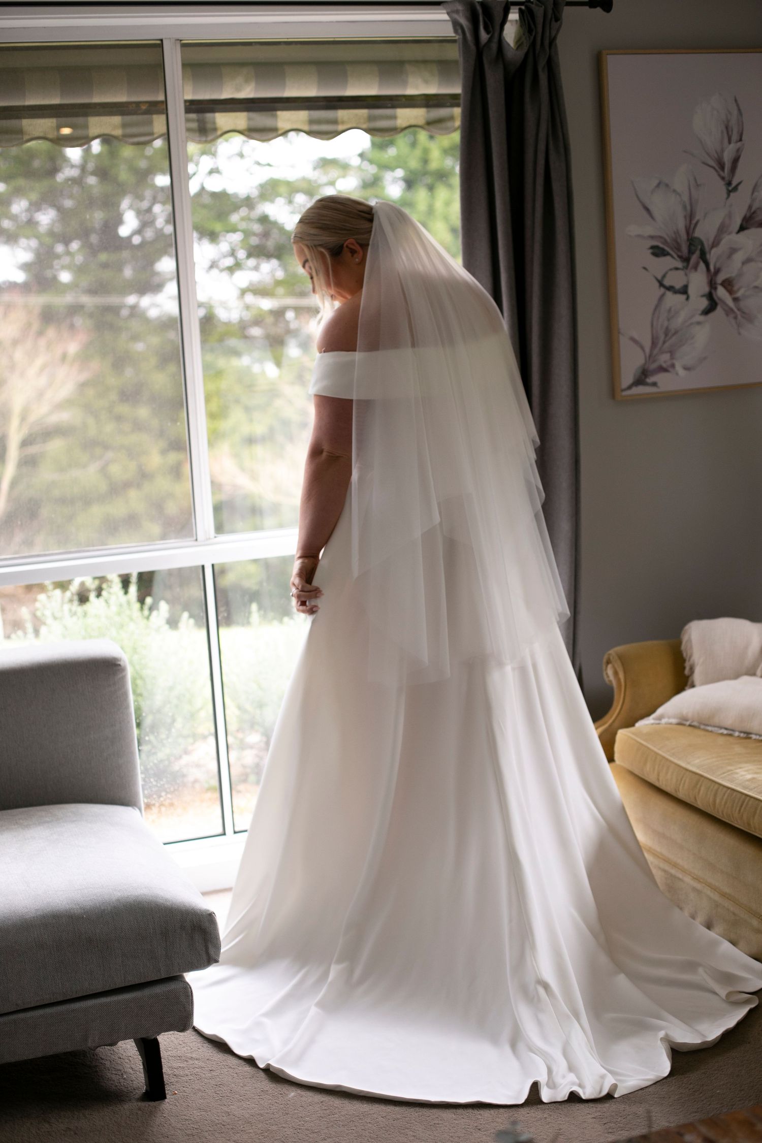 An elegant off-shoulder wedding gown with a flowing train and veil is showcased near a window in natural light.