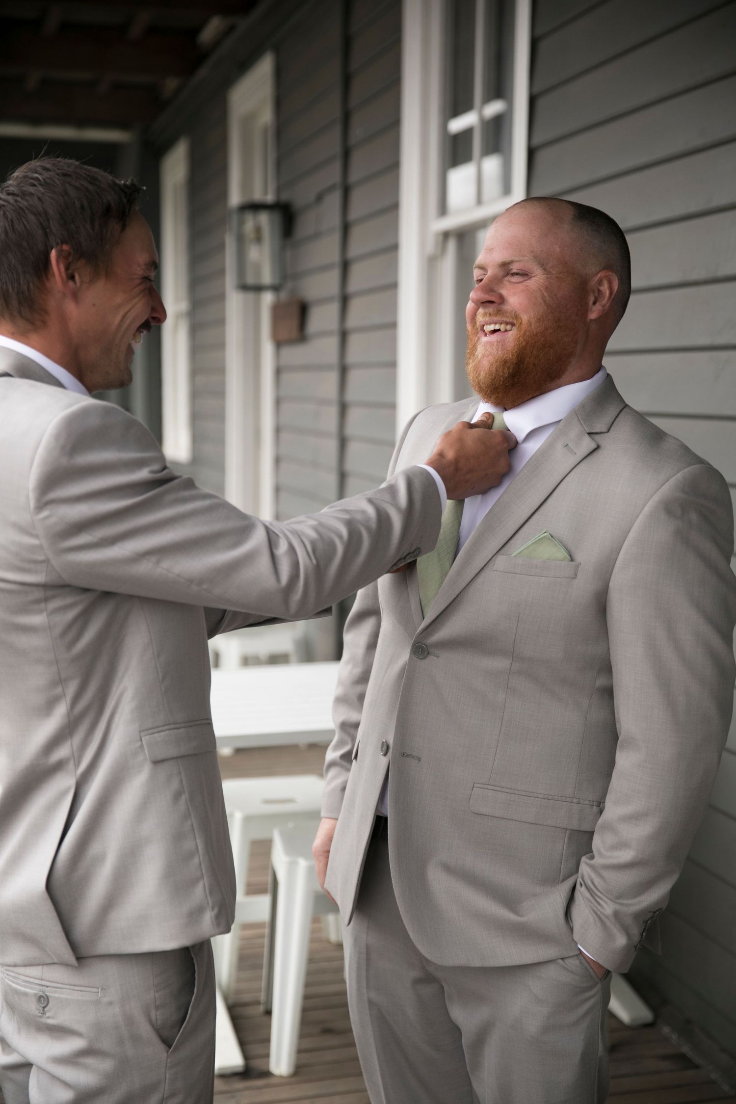 Two men in matching light gray suits share a lighthearted moment on a porch, adjusting each other's formal attire.