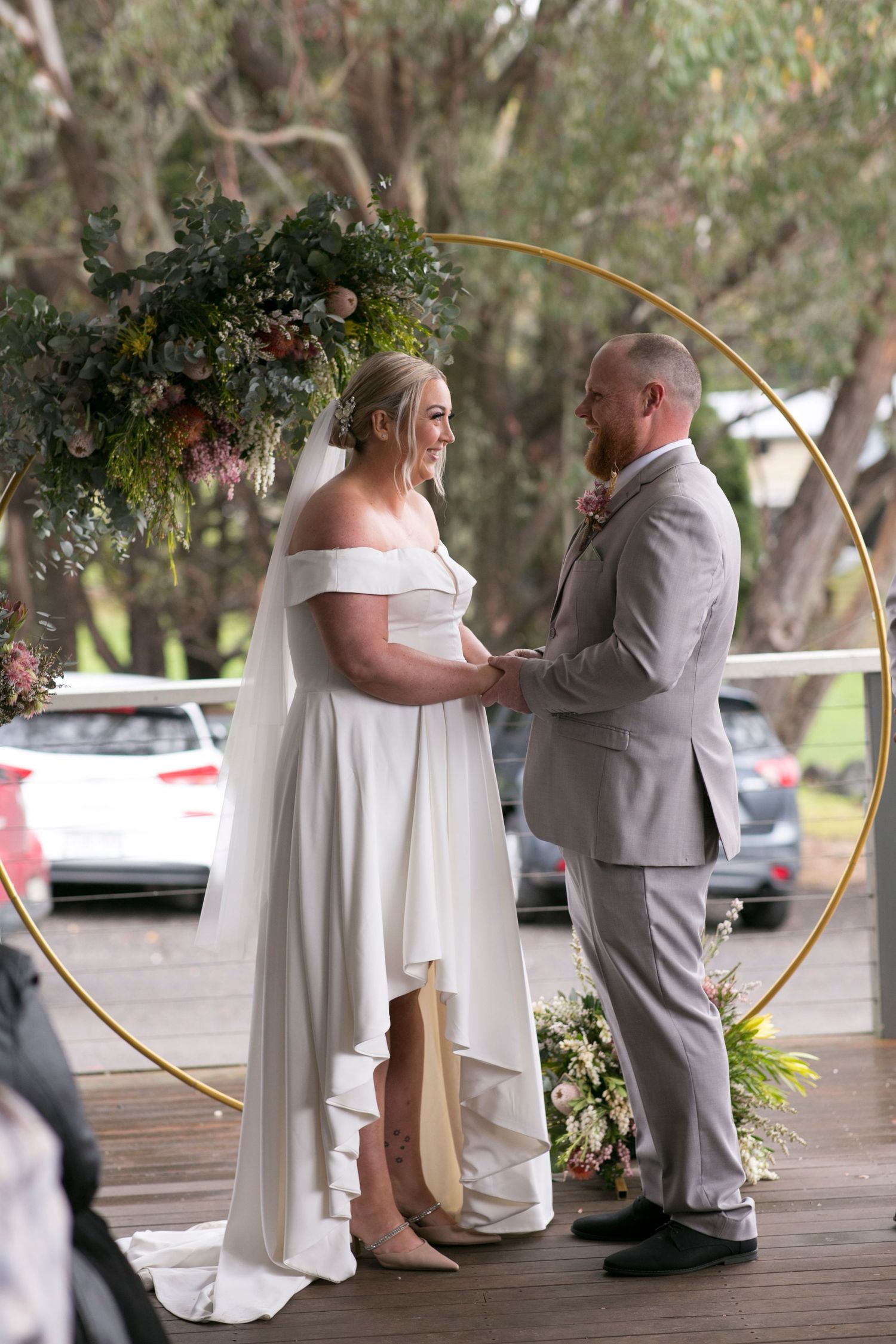 A couple stands together during their wedding ceremony under a circular floral arch on a wooden deck.