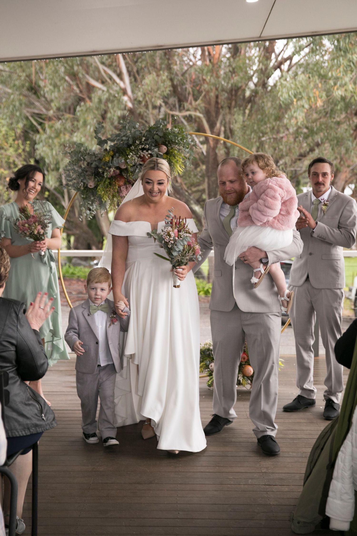 A wedding party walks down a wooden deck outdoors featuring a bride in white and wedding party in grey and mint green.