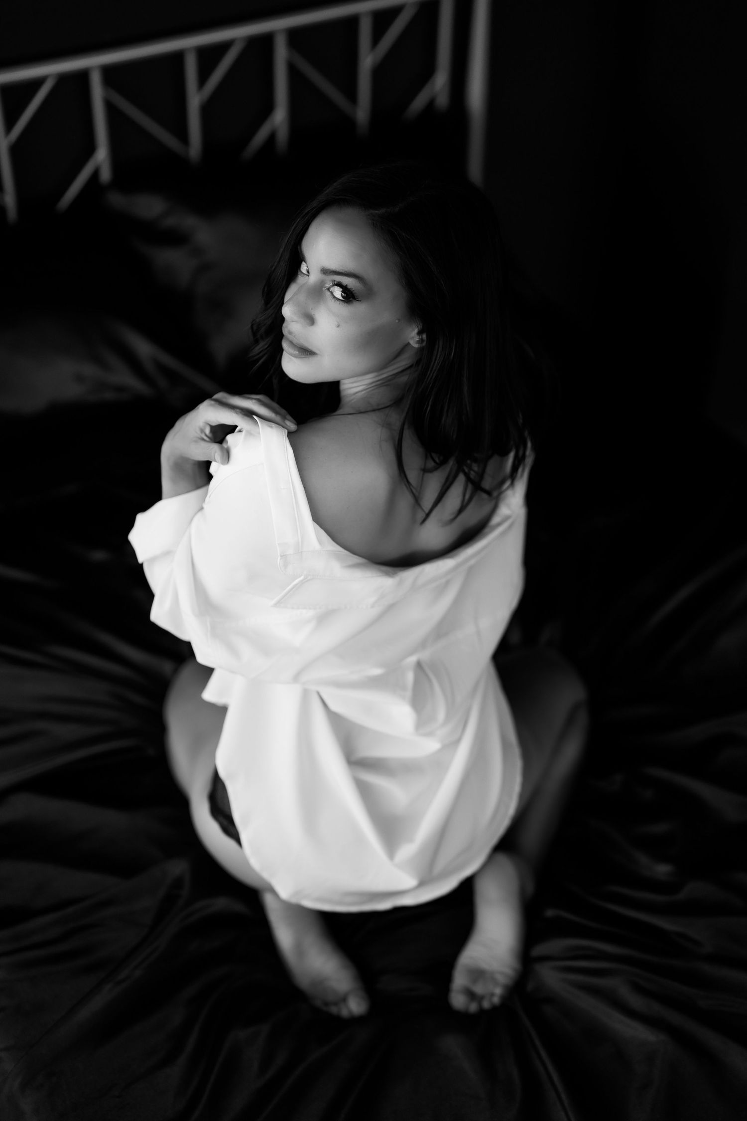 Dramatic black and white boudoir photograph of someone reclining on a bed near a window with soft natural light.