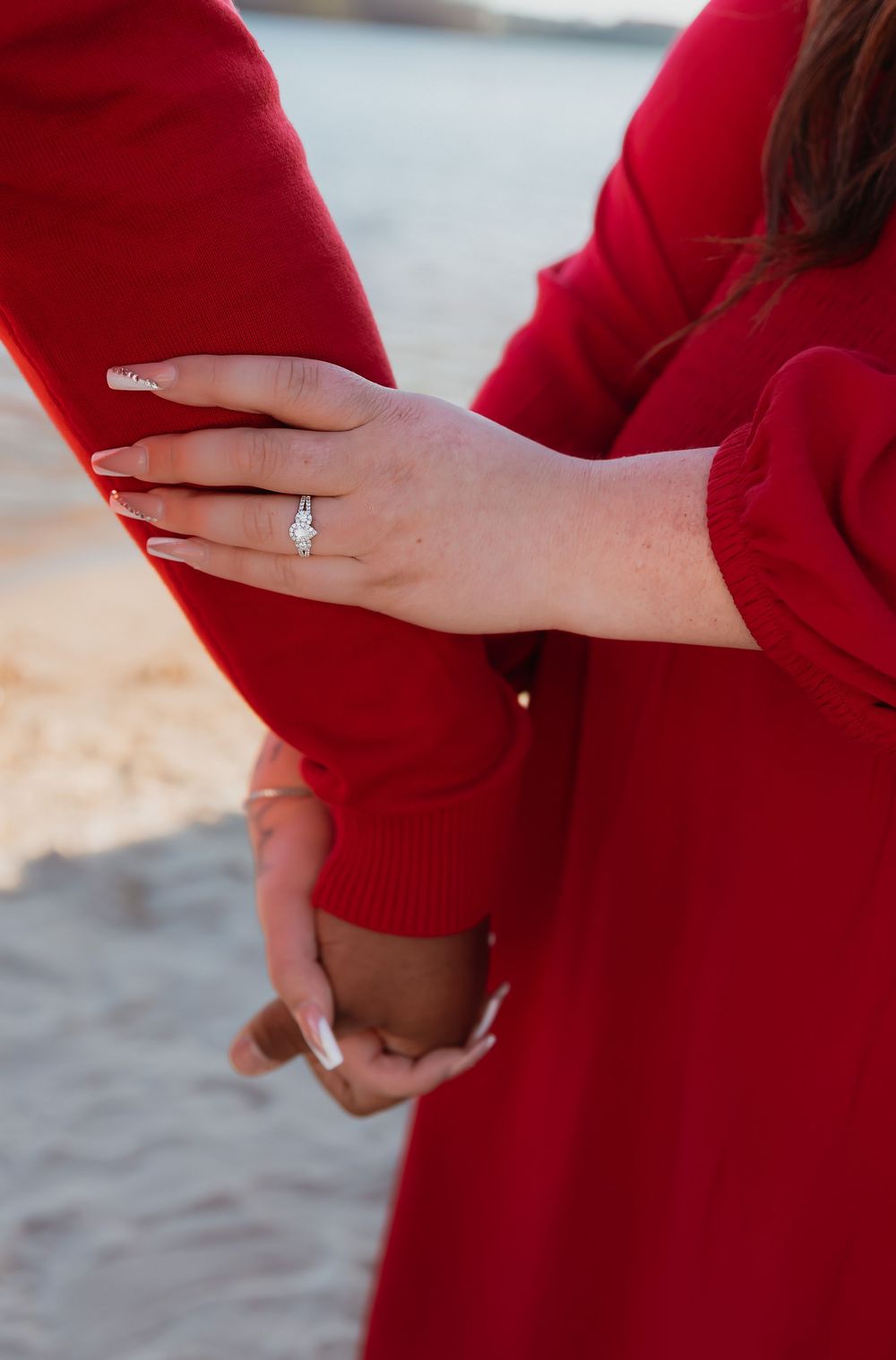 Person in a red sweater showing off an engagement ring while sitting on sand at sunset.
