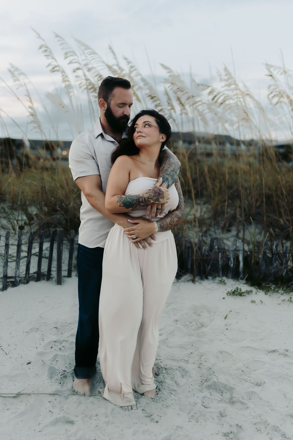 A couple embraces on a sandy beach with sea oats swaying in the background at dusk.