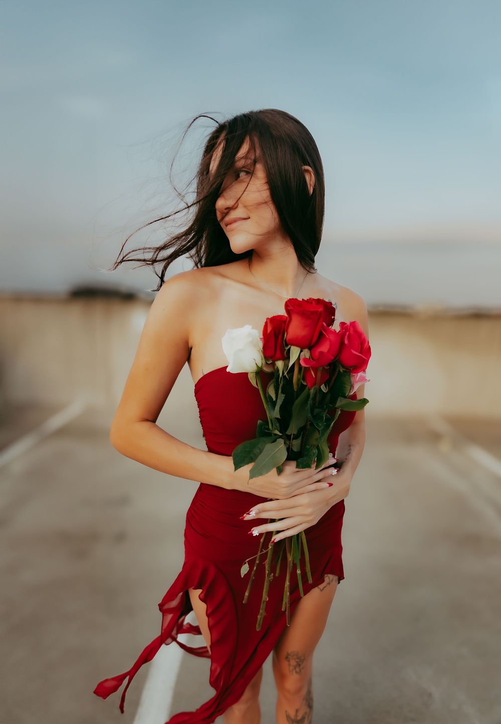 Woman in a flowing red dress holds vibrant red roses against a dreamy blue sky backdrop.