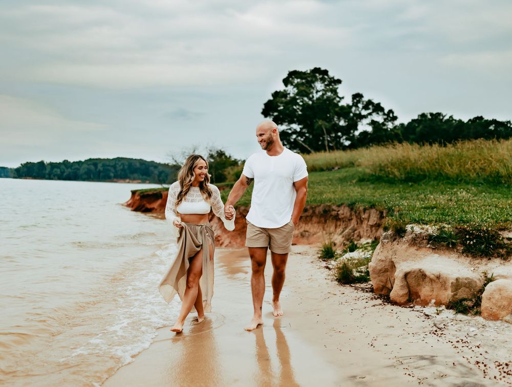 A couple walking together on a sandy beach shoreline with trees and water in the background on a hazy day.