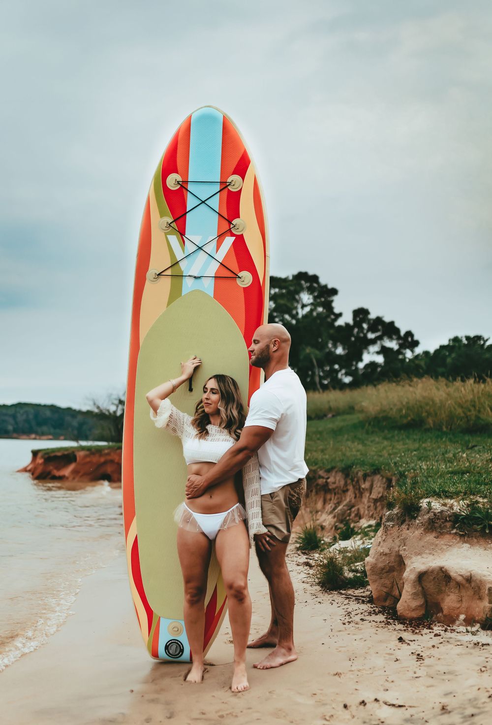 A couple poses on a beach with a red and blue paddleboard against a moody sky background.