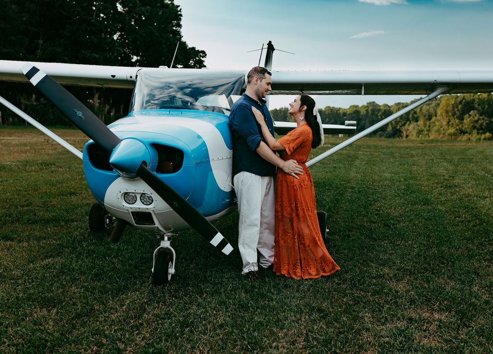 A couple poses together next to a small blue and white Cessna airplane on a grassy airfield.
