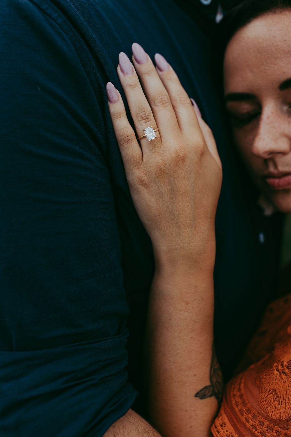 Close up of an engagement ring on a hand with purple manicured nails against a dark blue background.