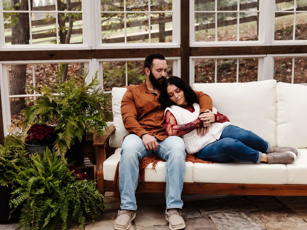 A couple relaxes together on a white cushioned bench surrounded by lush ferns in a sunlit greenhouse setting.