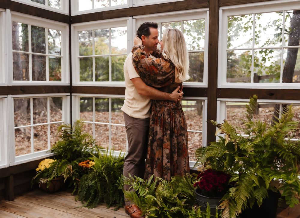 Two people share a romantic moment in a sunlit conservatory surrounded by lush greenery and potted plants.