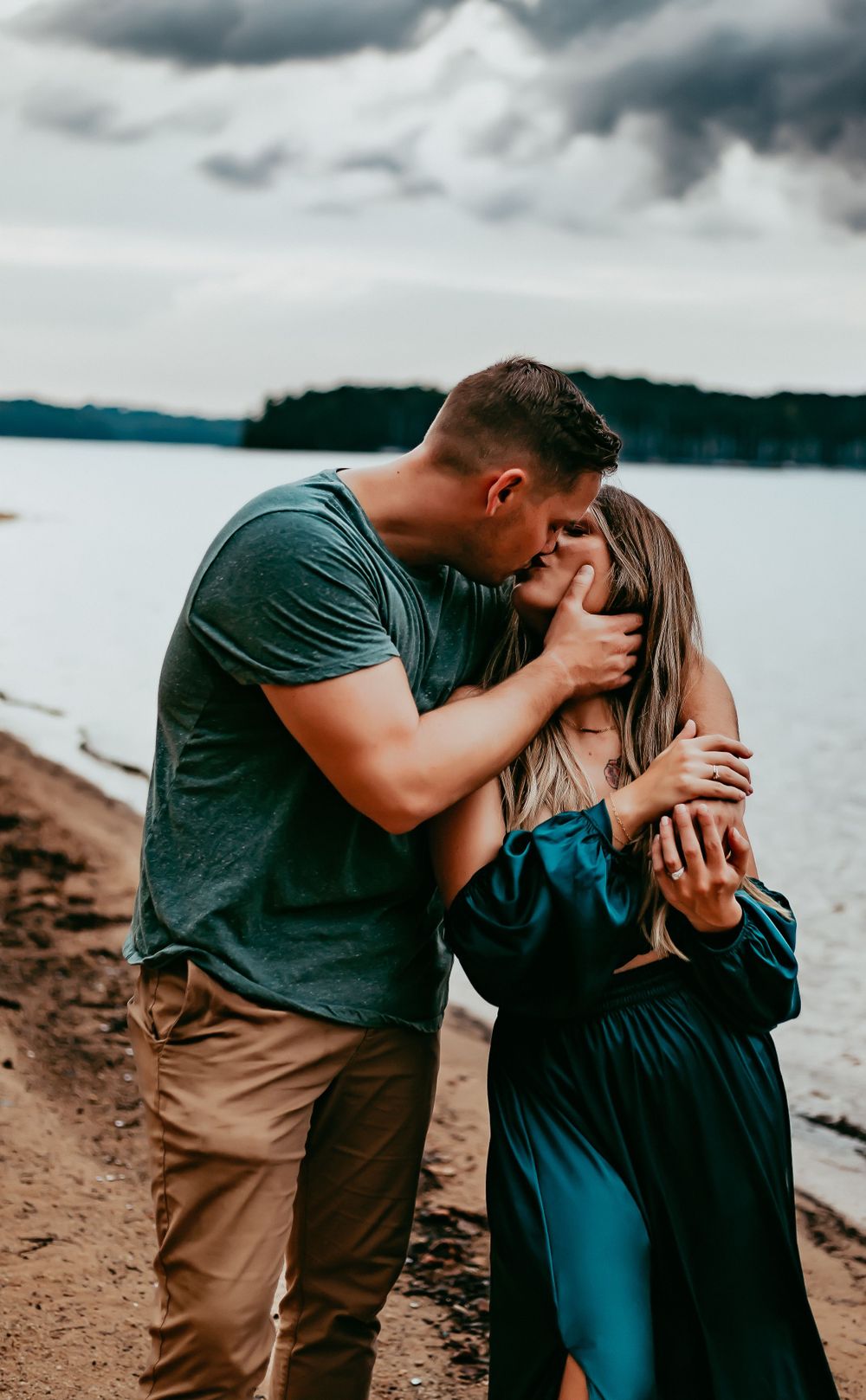 Romantic couple sharing a tender moment on a cloudy beach shoreline with water and trees in background.