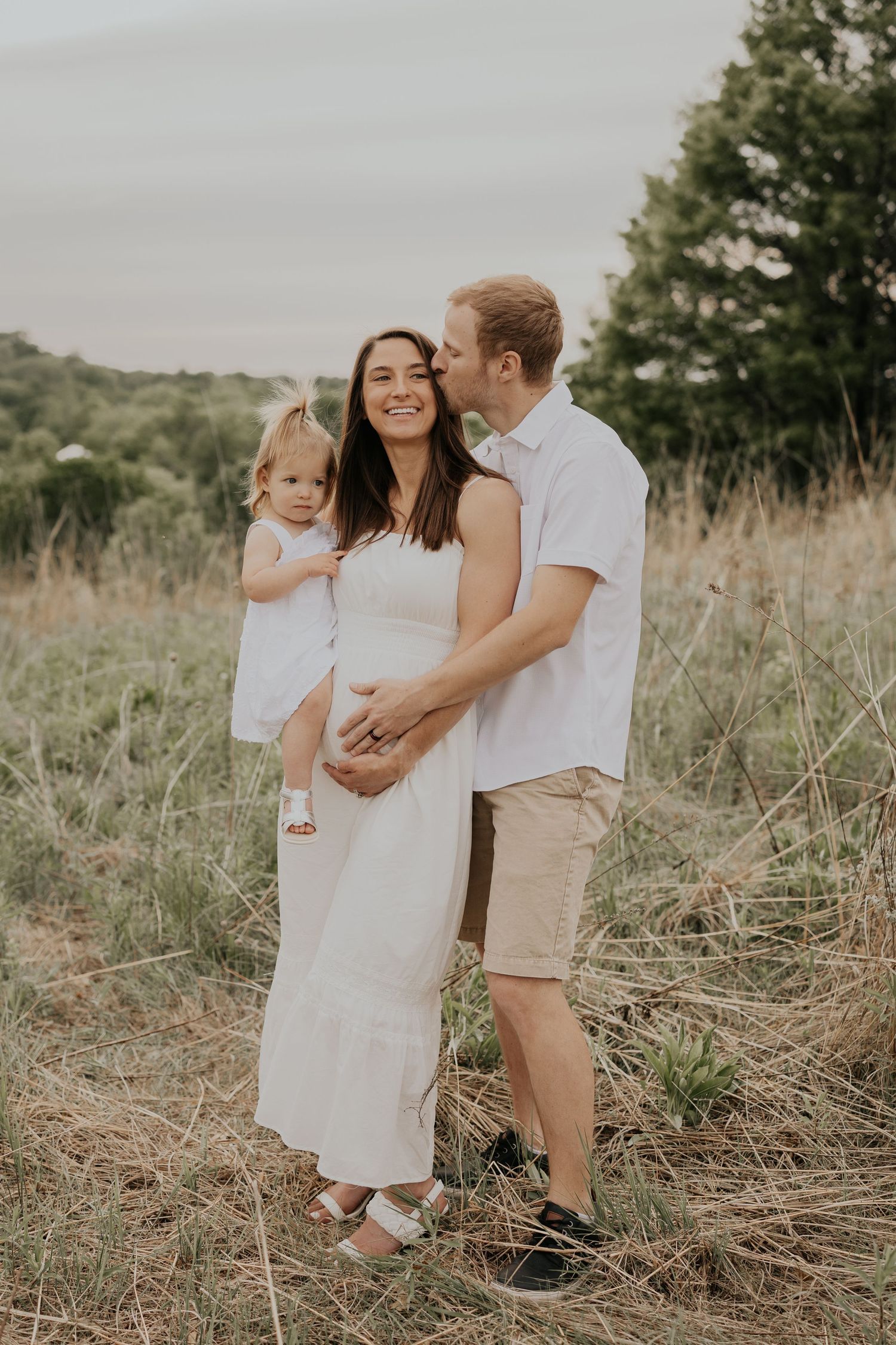 Family of three poses together in a grassy field wearing white and neutral colors for an outdoor portrait session.