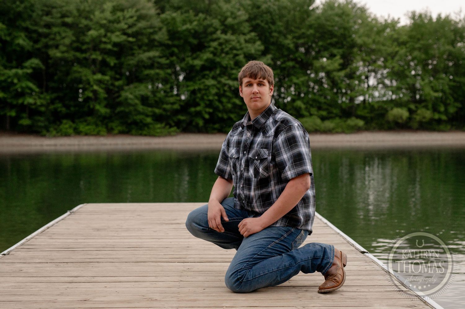 Person sits casually on wooden dock overlooking tranquil lake surrounded by green trees on summer afternoon.