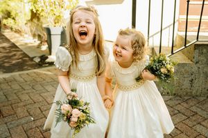 Two flower girls in white dresses laugh joyfully while holding bouquets on a brick patio.
