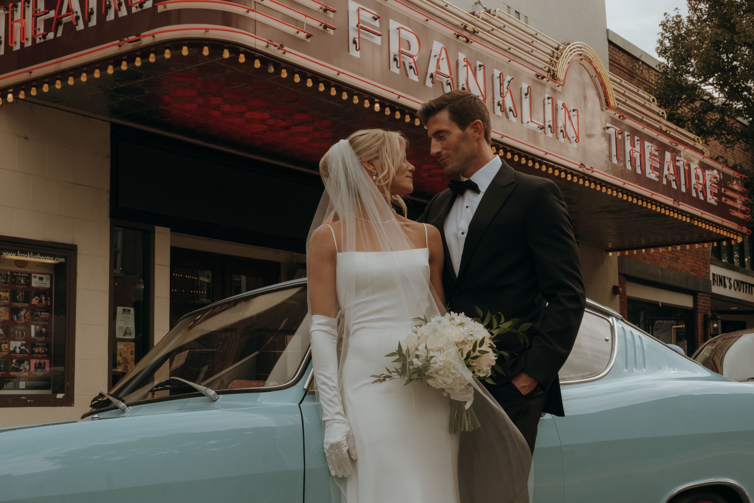 Elegant wedding couple sharing a romantic moment outside a vintage theater with glowing marquee lights at sunset.