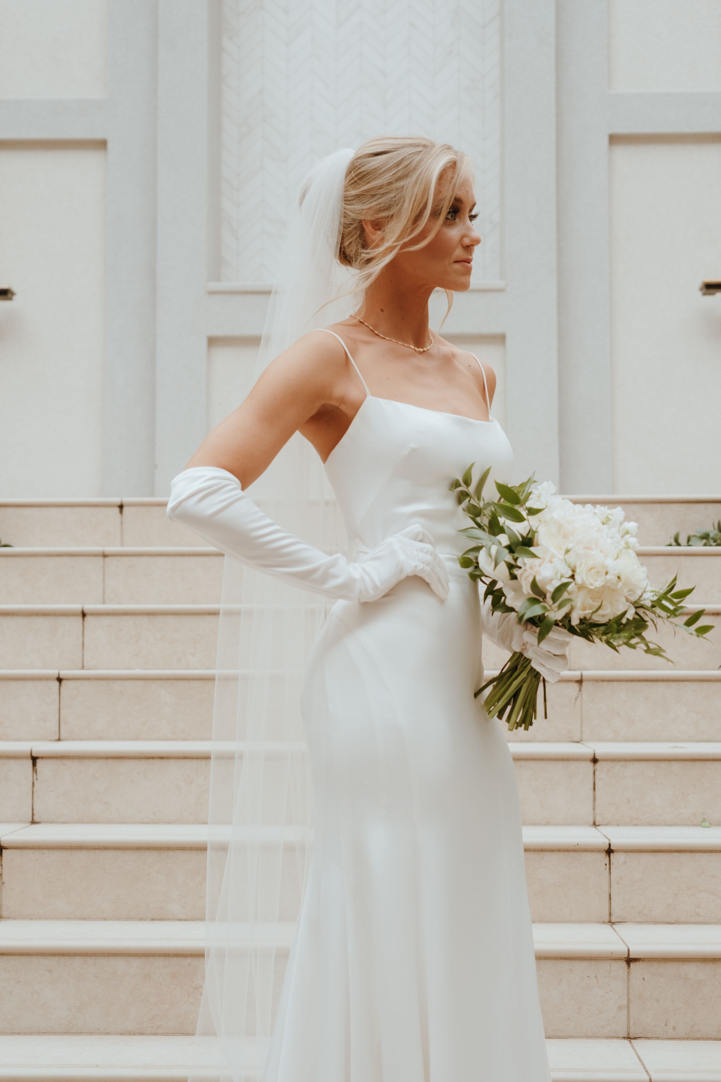 Elegant bride in sleek white satin gown and long gloves holds a white bouquet while standing on stone steps.