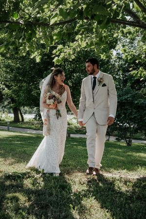 Newlyweds walking hand in hand through a lush green garden path in summer light.