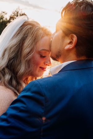 A bride and groom share an intimate moment in warm sunset light, with her white veil glowing against his navy blue suit.