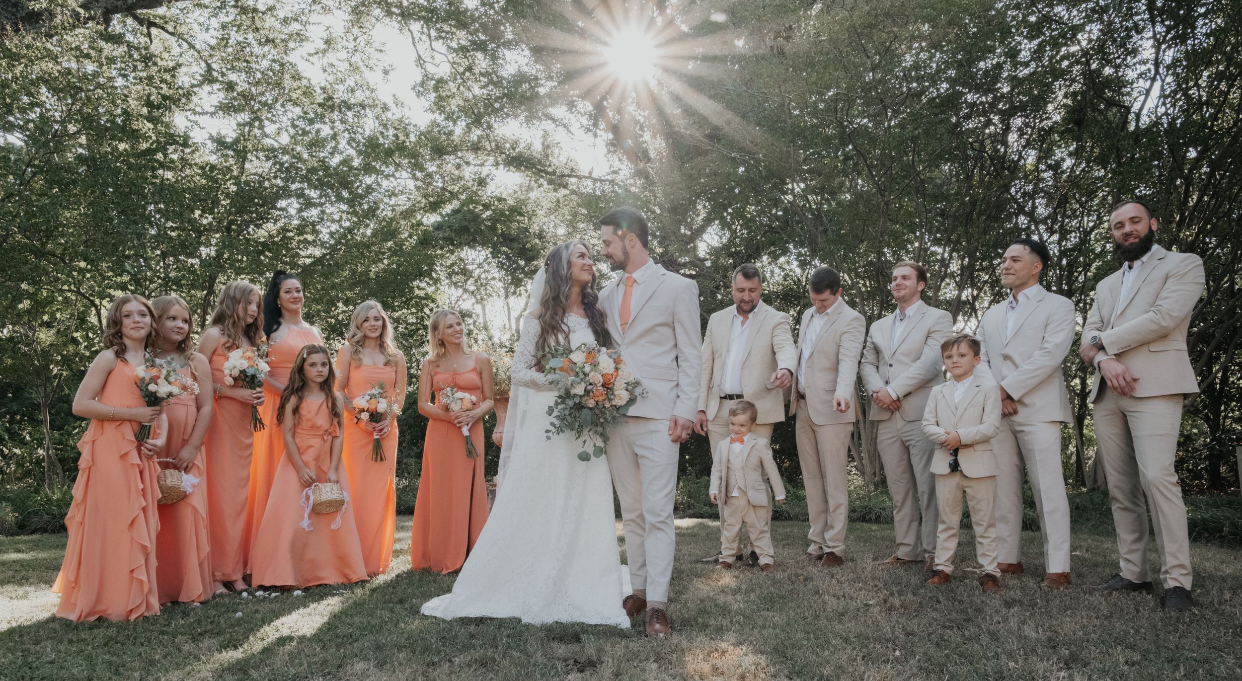 A wedding party in peach and neutral colors poses together on a scenic outdoor lawn at sunset.