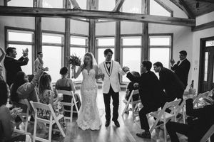 Wedding ceremony guests seated in white chairs inside a rustic venue with exposed wooden beams.