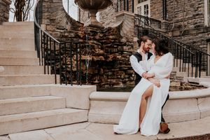 Couple in wedding attire embracing on stone steps against a rustic brick wall.