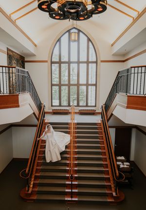 Dramatic view of grand staircase with arched window and white wedding dress flowing down carpeted steps.