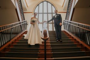 A couple stands on opposite sides of an elegant staircase with wooden railings and a large arched window behind them.